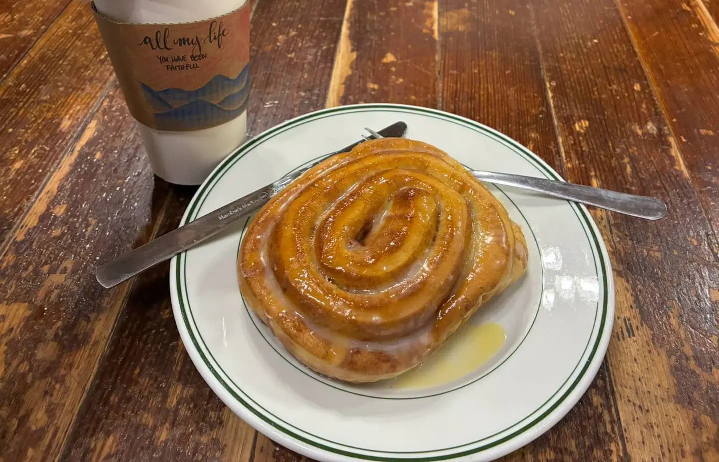 A glazed cinnamon roll on a green-rimmed plate beside a coffee cup on a wooden table